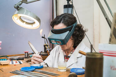 Seitenansicht einer konzentrierten reifen Meisterin mit Brille bei der Herstellung von Schmuck in der Werkstatt - ADSF20257