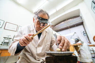 Niedriger Winkel des fokussierten reifen männlichen Meisters mit Brille, der Hammer und Meißel bei der Bearbeitung von Edelsteinen in einer Schmuckwerkstatt benutzt - ADSF20260