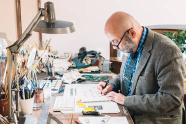 Seitenansicht des konzentrierten kreativen reifen männlichen Designers, der an Schmuckdesigns arbeitet und Skizzen zeichnet, während er an einem Tisch mit professionellem Zubehör am Arbeitsplatz sitzt - ADSF20261