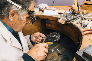 Hohe Winkel Seitenansicht der qualifizierten reifen männlichen Meister in Brillen mit Lötkolben bei der Arbeit an der Werkbank mit professionellen Instrumenten in Juwelier Werkstatt - ADSF20263