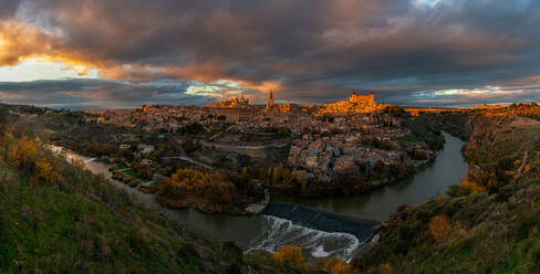 View across river of old city Toledo in Spain with medieval castles and fortresses at sunset time with cloudy sky and reflection in river water - ADSF20273
