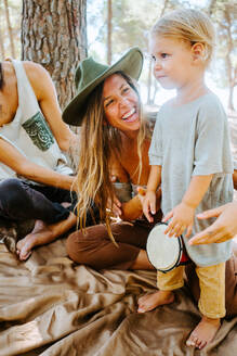 Delighted mother and crop father teaching little kid playing bongo drum while spending time together in woods - ADSF20283