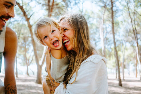 Multiethnische Mutter und Vater, die ihr kleines Kind zärtlich in die Wange kuscheln, während sie sich gemeinsam im Wald ausruhen - ADSF20285