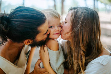 Side view of multiethnic mother and father cuddling and tenderly kissing little child in cheek while resting together in forest - ADSF20287