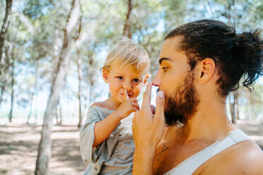 Side view of loving ethnic father gently cuddling cute child in cheek while standing in woods together at weekend - ADSF20294