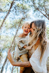 From below side view of positive mother with adorable little kid having fun and laughing together in woods during summer weekend - ADSF20296