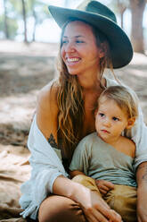 Positive mother with adorable little kid having fun and laughing together in woods during summer weekend - ADSF20301