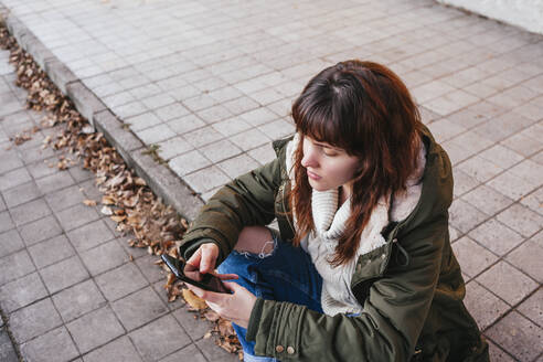 Junge Frau benutzt ihr Smartphone, während sie im Winter auf dem Gehweg sitzt - MGRF00147