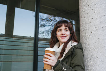 Smiling young woman holding reusable cup while looking away against column - MGRF00149