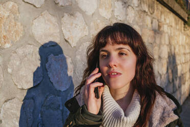 Contemplating young woman talking on smart phone against stone wall during sunset - MGRF00150