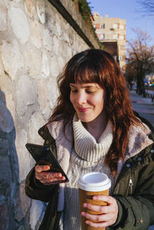 Smiling young woman using smart phone while holding reusable cup by stone wall - MGRF00151