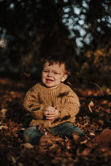 Nettes lächelndes Kleinkind sitzt inmitten trockener Herbstblätter im Wald bei Sonnenuntergang - GMLF00937