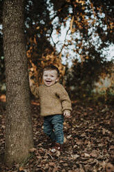 Cheerful toddler standing by tree trunk in forest during sunset - GMLF00940