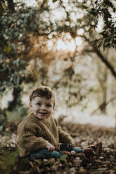 Cheerful toddler sitting amidst dry autumn leaves while looking away in forest - GMLF00941