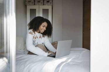 Smiling woman using laptop while sitting on bed at home - TCEF01464