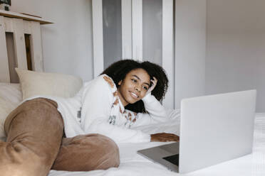 Smiling woman using laptop while lying on bed at home - TCEF01465