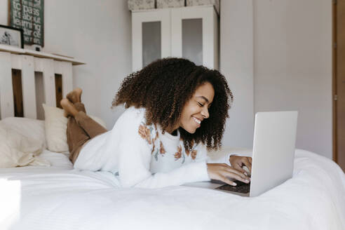Junge Frau lächelt, während sie einen Laptop benutzt und zu Hause auf dem Bett liegt - TCEF01466