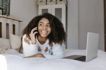 Glückliche Frau, die mit ihrem Handy telefoniert, während sie sich zu Hause auf dem Bett ausruht - TCEF01468