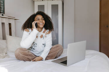 Lächelnde Frau mit Laptop, die zu Hause sitzt und mit ihrem Handy telefoniert - TCEF01469