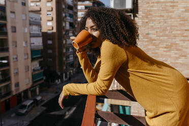 Woman drinking coffee while leaning on railing at balcony - TCEF01486