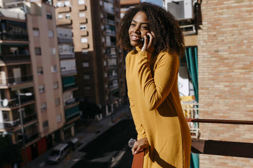 Frau mit lockigem Haar, die auf dem Balkon stehend mit ihrem Handy telefoniert - TCEF01487