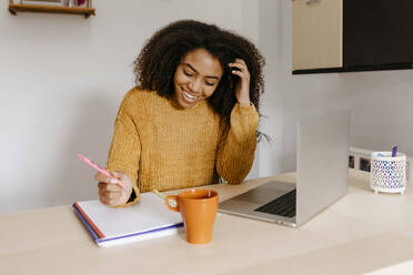 Smiling woman working over document while sitting with laptop at home office - TCEF01496