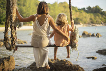 Mother and daughter on large rope swing over ocean, Thailand - FSIF05582