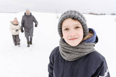 Junge, der in einer verschneiten Landschaft steht, mit Familie im Hintergrund - EYAF01461