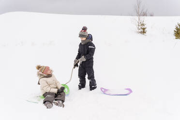 Brother and sister playing with sled on snow covered landscape - EYAF01464