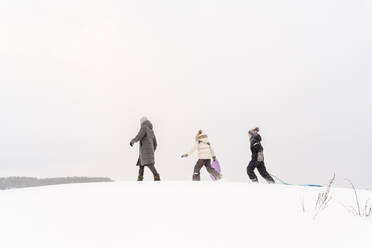 Father and children walking with sleds on snow covered land against sky - EYAF01470
