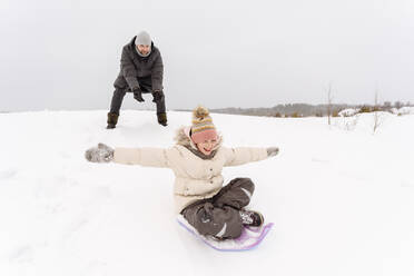 Father looking at cheerful daughter sledding on snow covered land - EYAF01471