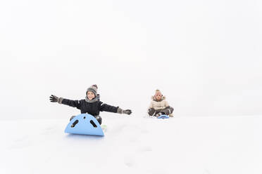 Playful sister and brother sledding on snow covered hill - EYAF01474