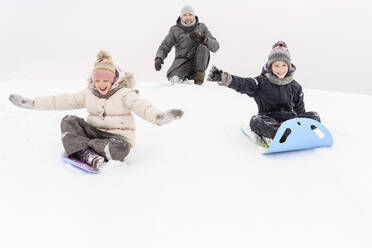 Vater betrachtet Kinder beim Schlittenfahren auf einem schneebedeckten Hügel - EYAF01475