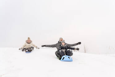 Verspielter Vater mit Kindern beim Schlittenfahren auf einem verschneiten Hügel gegen den Himmel - EYAF01476