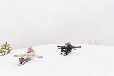 Playful father with children sledding on snowy hill against sky during vacation - EYAF01477