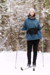 Lächelnde mittelgroße erwachsene Frau beim Skifahren auf schneebedecktem Gelände vor Bäumen im Wald - EYAF01488