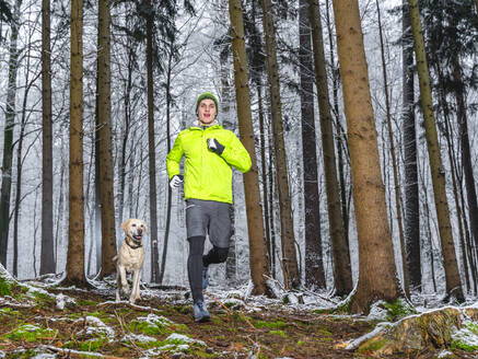 Männlicher Sportler joggt im Schnee mit Labrador Retriever im Wald - STSF02784