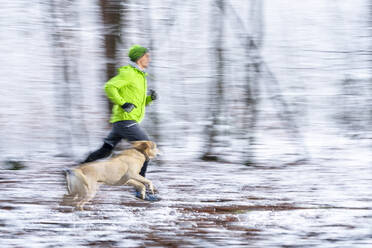 Young male athlete running in speed with dog during winter in forest - STSF02787
