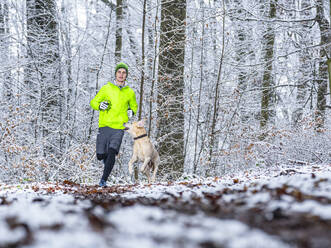 Young male athlete jogging with Labrador retriever during winter in forest - STSF02789