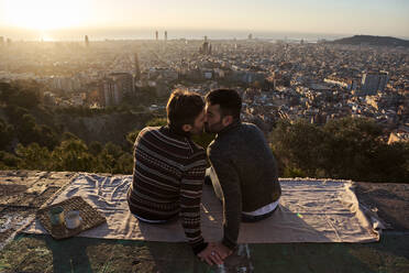 Schwule Männer, die sich küssen, während sie auf dem Aussichtspunkt vor der Stadtkulisse sitzen, Bunkers del Carmel, Barcelona, Spanien - VEGF03624