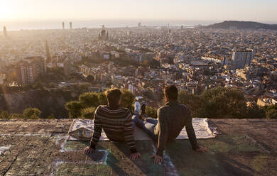 Gay boyfriends looking at crowded cityscape while sitting on viewpoint, Bunkers del Carmel, Barcelona, Spain - VEGF03629
