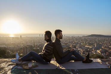 Schwules Paar sitzt Rücken an Rücken auf dem Beobachtungspunkt während des Sonnenaufgangs, Bunkers del Carmel, Barcelona, Spanien - VEGF03631