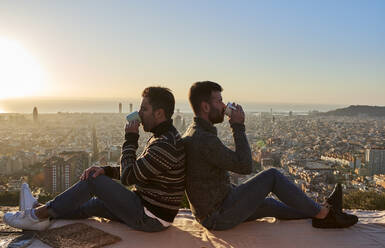 Ein schwules Paar trinkt Kaffee, während es Rücken an Rücken auf dem Aussichtspunkt des Bunkers del Carmel sitzt, Barcelona, Spanien - VEGF03632