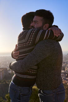 Verliebte schwule Männer, die sich umarmen, während sie bei Sonnenaufgang vor dem klaren Himmel stehen, Bunkers del Carmel, Barcelona, Spanien - VEGF03640