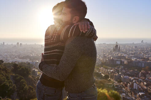 Freunde, die sich umarmen, während sie bei Sonnenaufgang vor einer Stadtlandschaft stehen, Bunkers del Carmel, Barcelona, Spanien - VEGF03642
