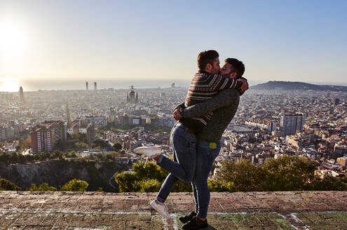 Schwule Männer, die sich küssen, während sie auf einem Aussichtspunkt vor dem Stadtbild stehen, Bunkers del Carmel, Barcelona, Spanien - VEGF03645