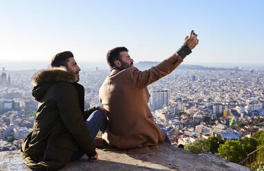 Mann macht Selfie mit seinem Freund, während er auf dem Aussichtspunkt sitzt, Bunkers del Carmel, Barcelona, Spanien - VEGF03649