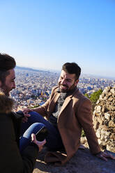 Glückliches homosexuelles Paar, das sich unterhält, während es auf dem Beobachtungspunkt gegen den klaren Himmel bei Bunkers del Carmel, Barcelona, Spanien, sitzt - VEGF03651
