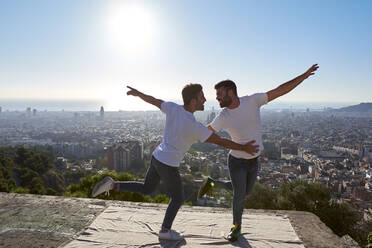 Glückliches schwules Paar mit ausgestreckten Armen tanzt auf dem Aussichtspunkt gegen den klaren Himmel, Bunkers del Carmel, Barcelona, Spanien - VEGF03658