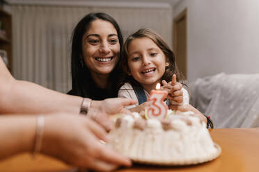 Smiling mother and daughter looking at birthday cake on table at home - EGAF01524